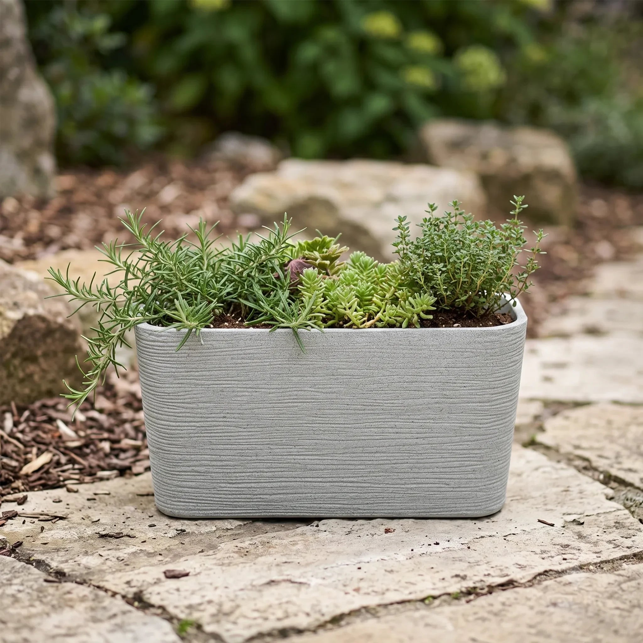 Nora 26-inch rectangular planter in a grey stone-look finish, filled with green herbs on a stone patio.