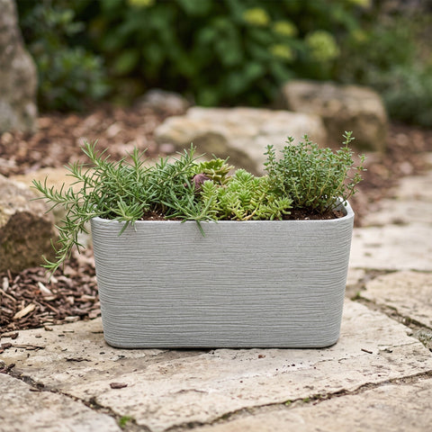 Nora 26-inch grey rectangular planter with a textured stone finish, filled with green herbs on a stone patio.