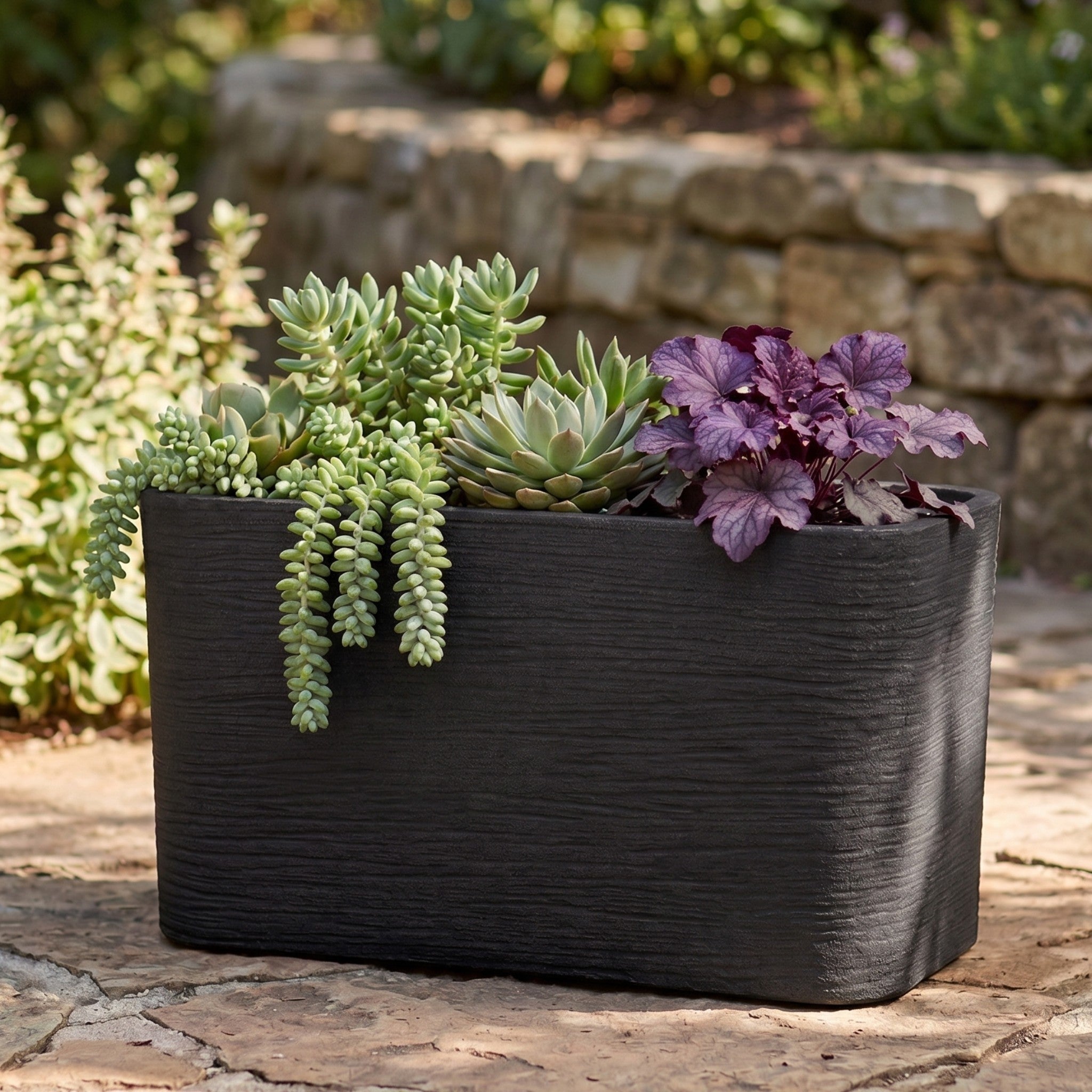 Nora 26-inch rectangular planter in a dark textured stone finish, filled with assorted succulents on a patio.